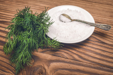 Fototapeta na wymiar Salt in the bowl and dill greenery on brown wooden table background.
