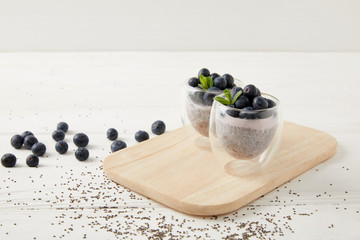 close up view of chia puddings with fresh blueberries and mint on wooden cutting board on white...