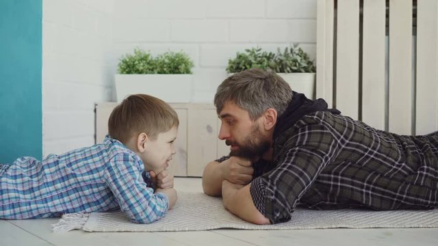 Cute small child is lying on floor with his loving dad, looking at him and talking. Happy family, interesting conversation, childhood and parenthood concept.