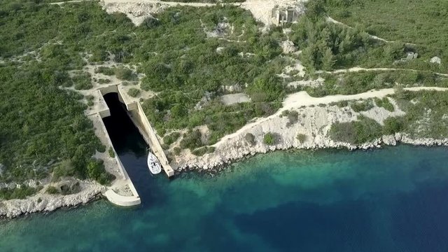Aerial: Flying in front of concrete naval abandoned submarine hiding hangar. Sailing crew using it to safely park their sailing boat in it.
