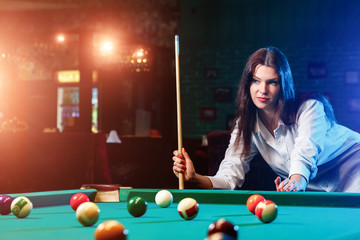 Young beautiful girl is playing billiards. Dark background. Pleasant pastime, rest, entertainment.