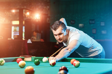 A man in a white T-shirt is playing billiards, a dark background. Pleasant pastime, entertainment, leisure, family holidays, games.