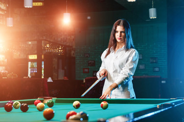 Young beautiful girl is playing billiards. Dark background. Pleasant pastime, rest, entertainment.