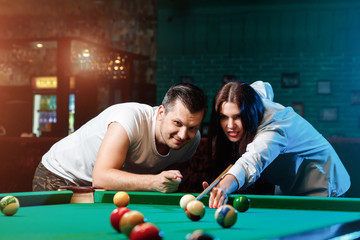 A guy and a beautiful girl are playing billiards, a guy is teaching a girl to play billiards. Dark background. Pleasant pastime, family rest, entertainment, holiday.