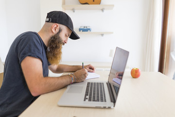 Fototapeta na wymiar Bearded young man sitting at desk with laptop writing down something