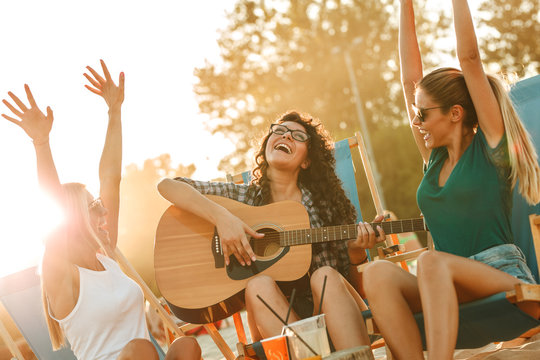 Group of young female friends sitting on beach on sunbeds,singing and playing guitar.