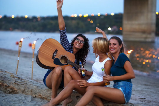 Group of young female friends sitting on the river beach ,singing and playing guitar.Joying in summer evening .