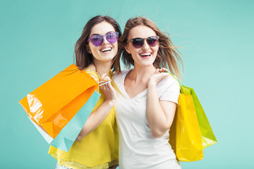 Two beautiful shopaholic smiling girls wears sunglasses holds lot of shopping colorful bags on blue background