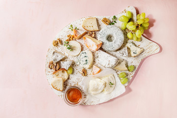 Cheese plate assortment of french cheese served with honey, walnuts, bread and grapes on white wooden serving board over pink pastel background. Top view, space.
