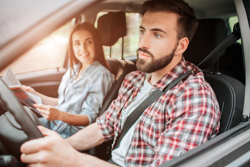 Careful and nice guy is driving car and looking straight forward. He is paying all of his attention to the road. Girl is sitting besides him. She is holding map and looking at young man with smile.