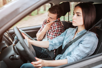 A picture of couple travelling in car. Guy is talking on the phone while girl is driving and paying attention to the road. She looks serious.