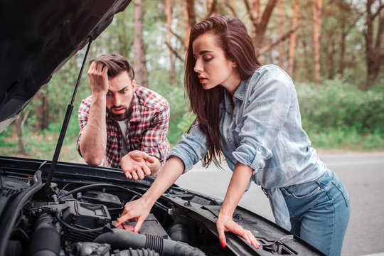A picture of girl trying to fix detail in car. She is leaning on car and looking to all detailes that are under hood. Guy is looking at all of this and wondering. He is tired and mad.