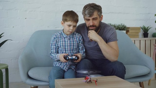 Cute child is holding transmitter and controlling flying helicopter while his father is trying to catch it with his hands. Family members are laughing and having fun.