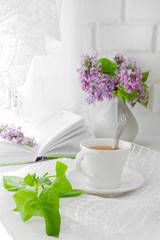 white cup with tea and a bouquet of lilac in a white vase on a summer morning breakfast at the window with a white lacy curtain in a rustic style with a copy of the space