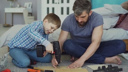 Little boy is learning to use electric screwdriver while his dad is explaining how to work with screw gun and fix screw in piece of wood. Construction and family concept.