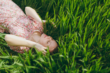 Young smiling tender beautiful woman with closed eyes in light patterned dress lying on grass resting in sunny weather in field on bright green background. Spring nature. Lifestyle, leisure concept.