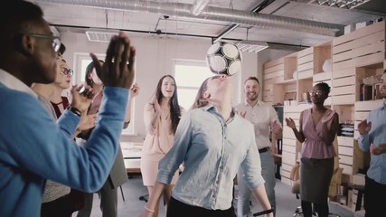 European girl juggling football on head in modern trendy office. Multiracial colleagues celebrate clapping, slow motion - Powered by Adobe