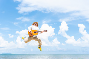 Chubby man wearing hat jumping while playing guitar along the beach. Happy man having a good mood jumping in the air.