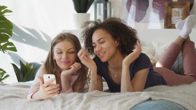 Two young attractive women caucasian blonde and brunette talk surprisingly on mobile Skype via webcam laugh in cozy bed in sunny room in Scandinavian style design with pastel coloured textile