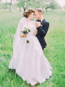 Vertical portrait of the happy newlywed couple standing head-to-head at the background of the green field.