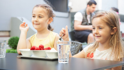 Obraz na płótnie Canvas two sweet girls in the restaurant eat red strawberries with cream.Two girls sharing bowl of fresh strawberries on beautiful spring day.