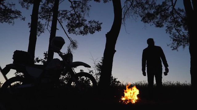 The silhouette of a cross off road motorcycle on a background of forest part in the evening, tent camp fire, bonfire, the man throws wood on the fire, and drinks tea, coffee from a cup