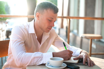 Man writing on notebook or planner with smartphone and coffee at outdoor cafe.