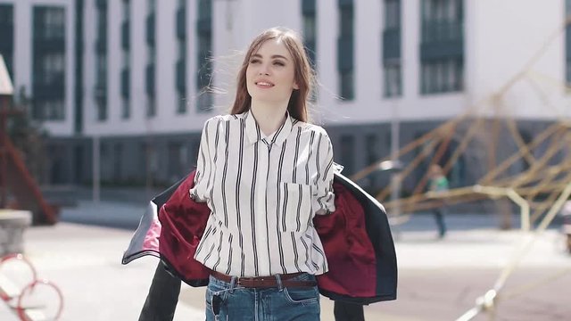 A young girl walks around the city and on the move takes off her jacket. portrait of a beautiful young woman against a background of modern architecture. slow motion