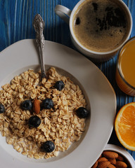 Oatmeal porridge with fresh blueberries, almonds, coffee and orange juice in white bowl on blue table. Healthy breakfast, healthy eating, vegan food concept.