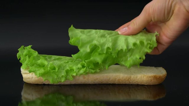 preparing fresh submarine sandwich with lettuce leaves and ham on black background