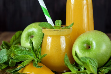 Yellow smoothie in glass jars surrounded by fruits and mint against wooden background