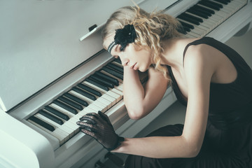 Young actress lying on piano overwhelmed with memories. Sad blond girl leaning on keyboard. Roaring twenties fashion concept