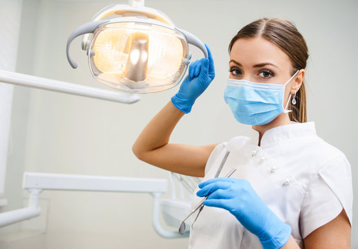 Young female friendly dentist in umiform standing in her office and looking at camera and smiling. Close up potrait of the doctor woman at white background