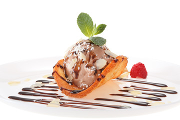 Chocolate ice cream ball decorated with chocolate syrup, nuts, raspberries, mint leaves and coconut chips on a wafer of coconut and honey. All on white plate isolated on white background.