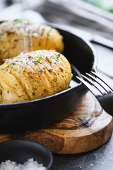 Homemade baked potato with olive oil, garlic, rosemary, mustard, dill and black pepper in a cost fry pan is being sprinkled sea salt.