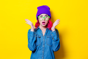 Young pink hair girl in blue shirt, purple hat and green glasses. Portrait isolated on yellow background