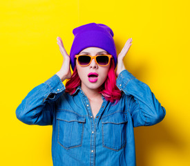 Young pink hair girl in blue shirt, purple hat and orange glasses. Portrait isolated on yellow background