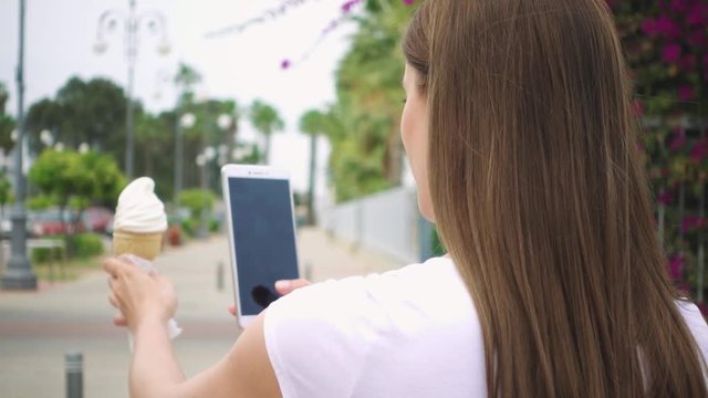 Young woman taking photo of ice cream cone outdoors in summer. Happy female teenager photographing vanilla ice cream on her camera phone on street in slow motion