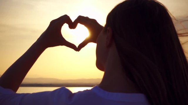 Dark silhouette of young woman at sunset on lake making heart gesture with fingers. View from behind of female figure at golden hour in slow motion catching sun in hands folded in heart shape