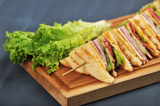 A sandwich with a filling of ham, bacon, tomato, pickled cucumber, roasted egg, cheese, sauce and salad. The sandwich is cut into four pieces and is put on a skewer. Close-up. Dark background.