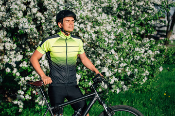 Professional cyclist in helmet and spotswear standing with mountain bicycle near blooming tree. Young rider resting in park after ride.