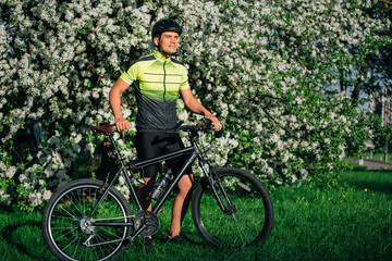 Professional cyclist in helmet and spotswear standing with mountain bicycle near blooming tree. Young rider resting in park after ride.