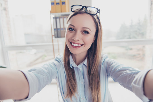 Self portrait of cheerful, positive, charming woman with hairstyle and glasses on head shooting selfie with two arms on front camera of smart phone in work place, station