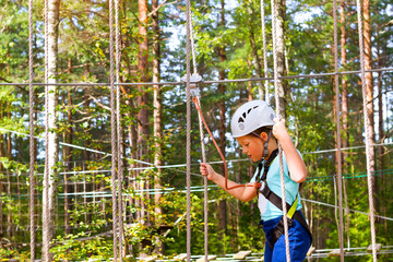 Teenage girl goes on hinged trail in extreme rope Park in summer forest. High-altitude climbing training of child on adventure track, equipped with safety straps and protective helmet. Estonian summer