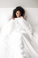 Fototapeta premium Portrait of beautiful smiling lady with dark curly hair lying in bed while dreamily looking in camera at home isolated