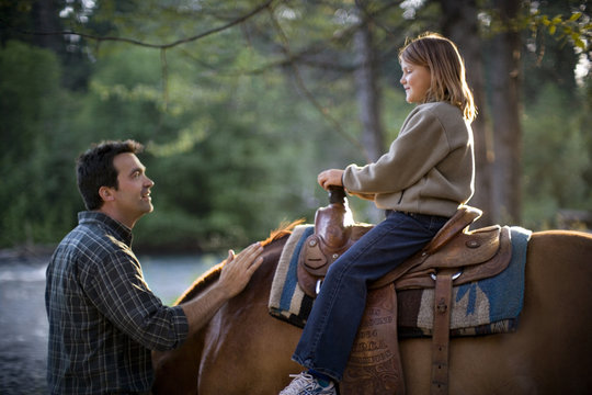 Mid-adult man talking to his teenage daughter atop a horse.