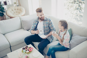 Portrait of stylish bearded trendy dad with red hair drinking beverage with little cute kid with hairstyle having sweets on the table holding mugs and saucers looking at each other enjoying talk