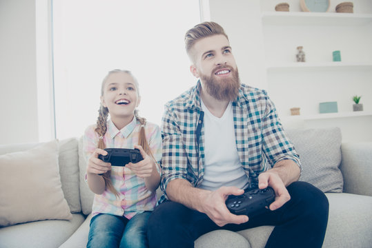 Cheerful joyful excited stylish trendy father and daughter holding joy-sticks in hands playing video game sitting on couch indoor in living room enjoying free time wearing casual outfit front view