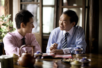Mature adult businessman sitting with a young adult male colleague drinking tea at a cafe.