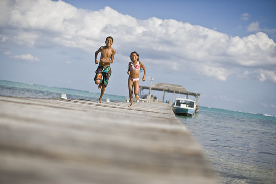 Smiling young girl and her older brother running along a wooden jetty in their swimwear.
